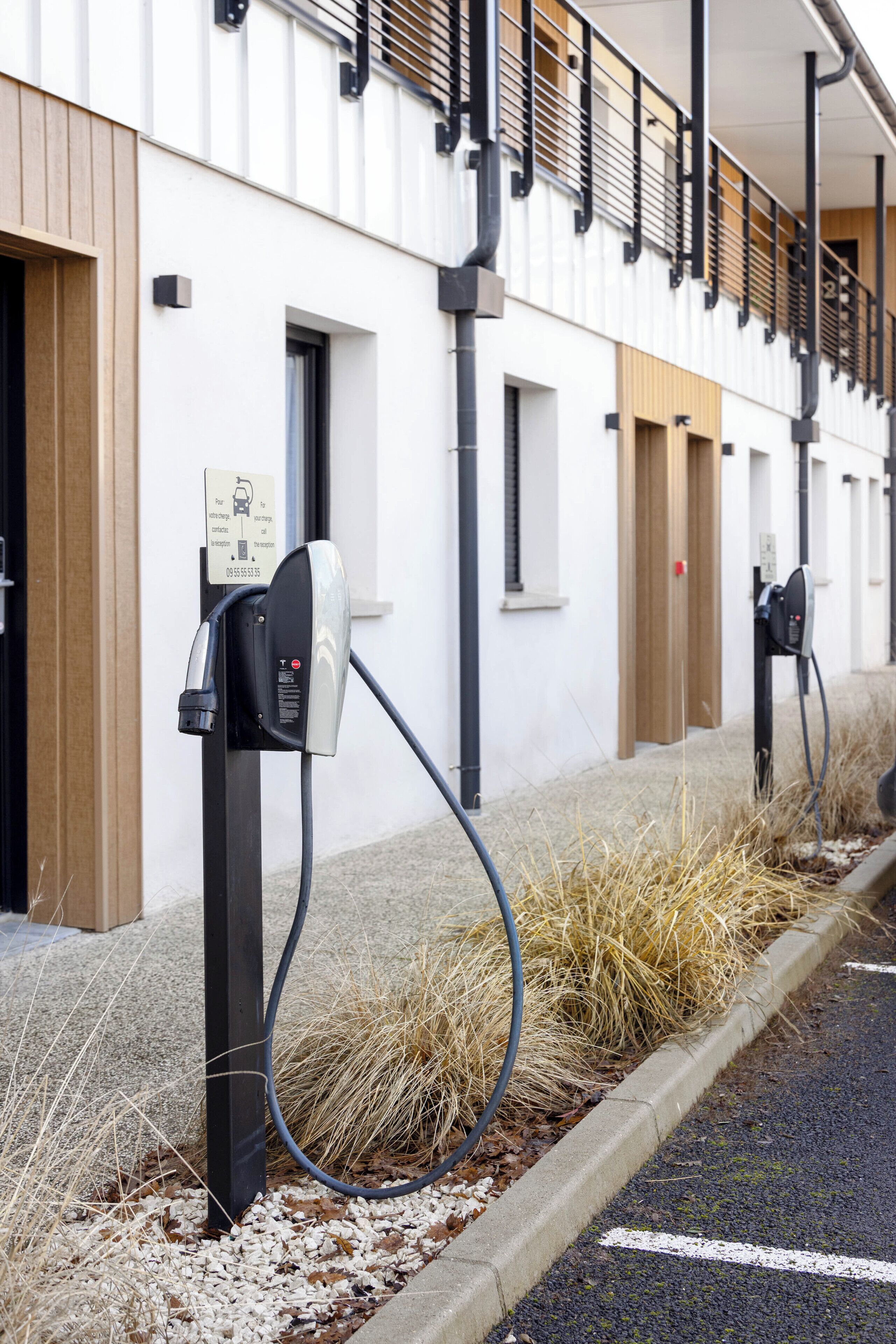 A parking area with electric vehicle charging stations, a building with a white facade, and a small garden bed with dry grass.