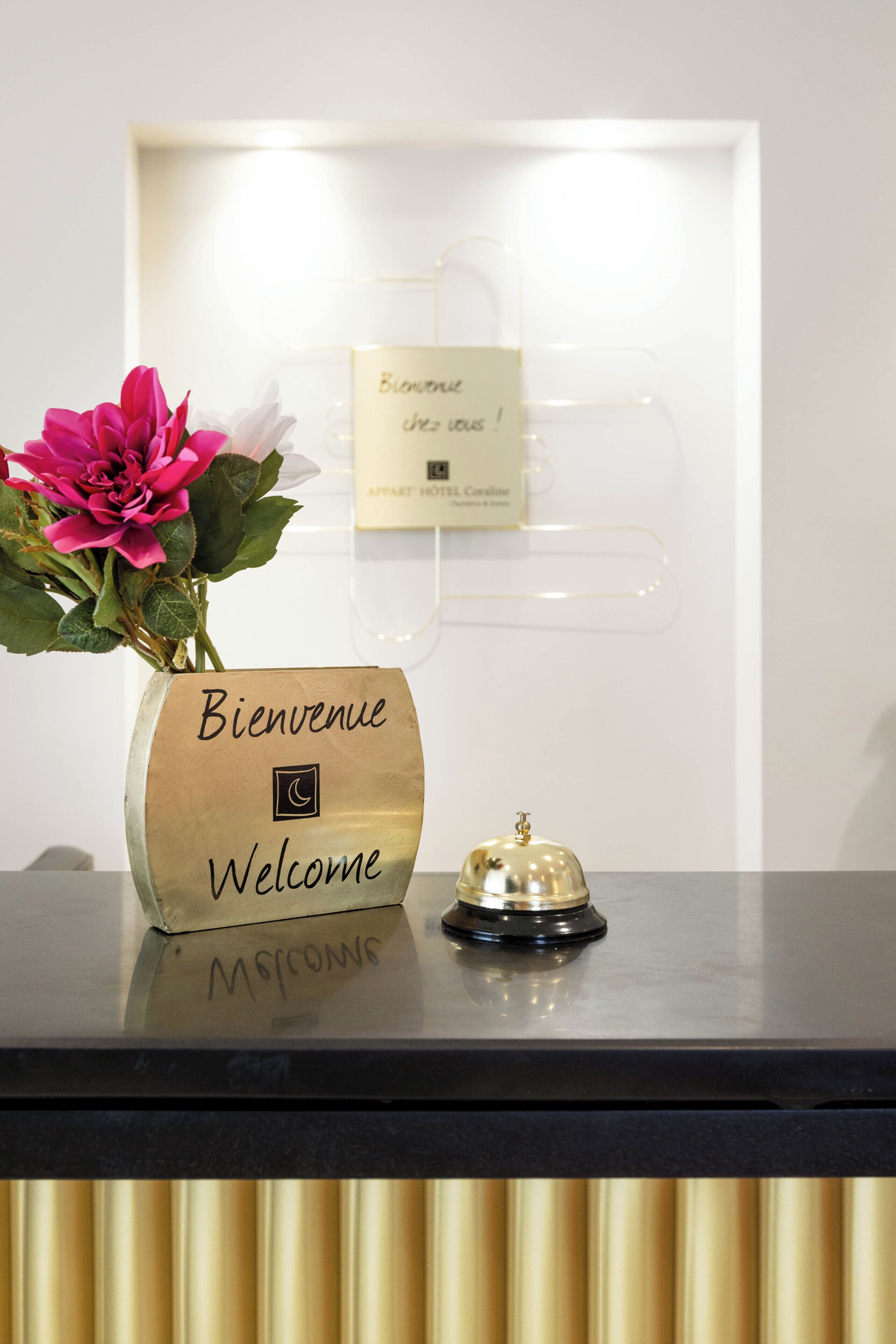 A reception desk with a 'Bienvenue Welcome' sign, a flower arrangement, a bell, and a card with a message.