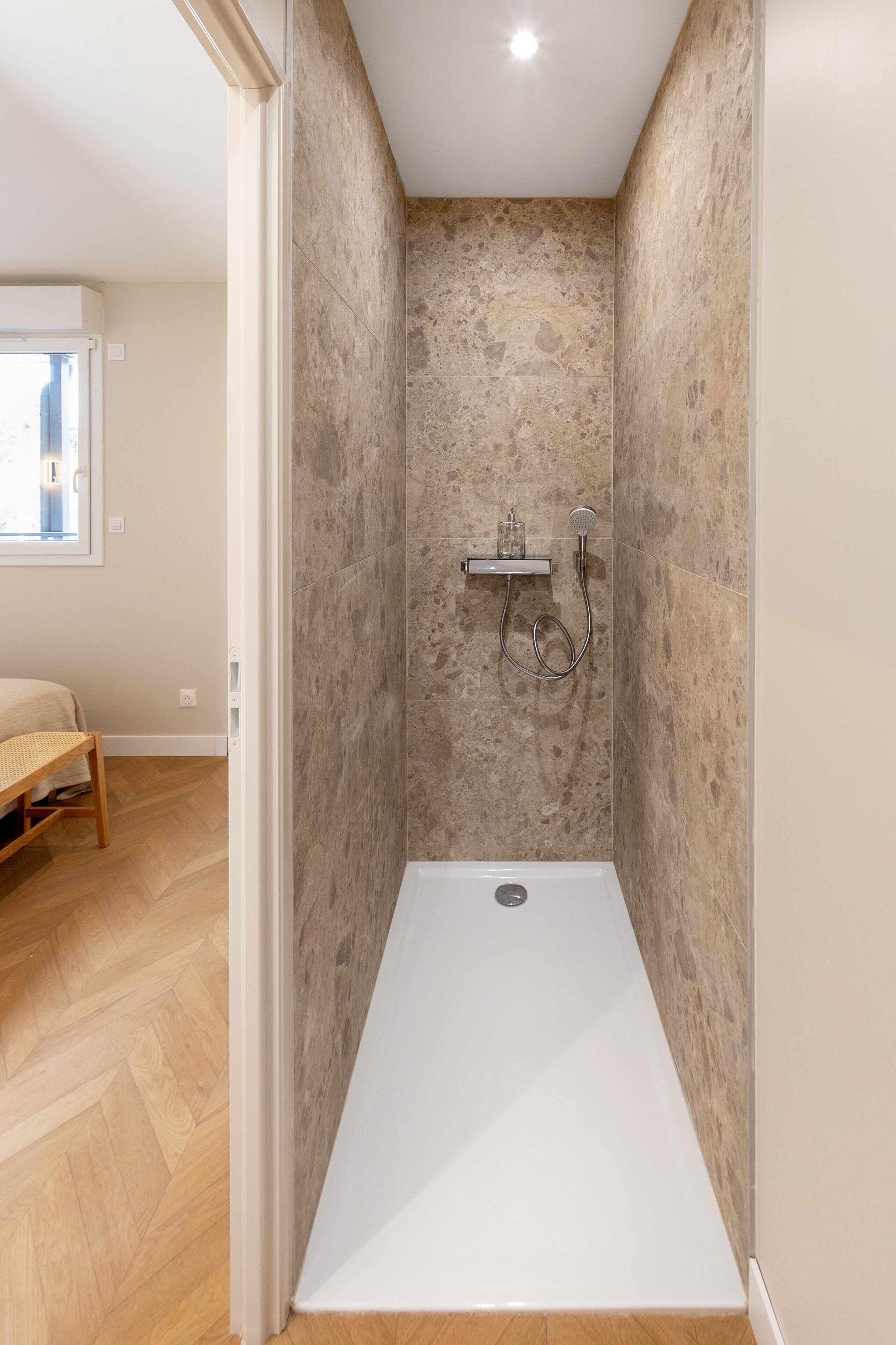A small, modern bathroom with a shower, a wooden bench, and a window.