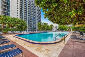 Piscina com espreguiçadeiras e palmeiras, cercada por edifícios altos.