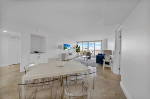 Uma sala de estar e jantar moderna e integrada, com uma grande mesa de jantar, cadeiras transparentes, área de cozinha e um espaço de estar com vista para o céu.