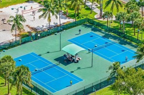 An outdoor tennis court with a green roof pavilion, surrounded by palm trees and a sandy area.