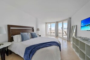 A hotel room with a large bed, a TV, a desk, and a balcony with a city view.