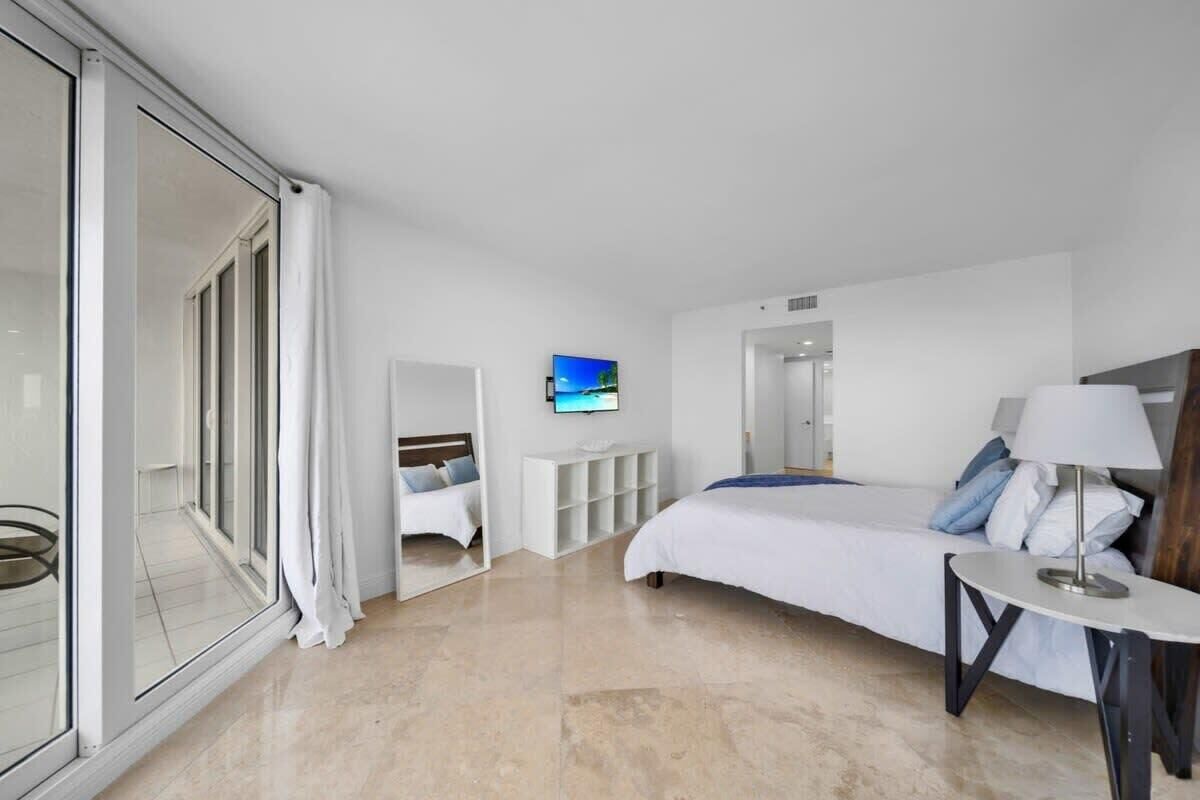 Ett modernt hotellrum med en stor säng, en tv som är monterad på väggen, ett litet bord och en skjutdörr i glas som leder ut till en balkong.