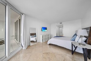 A modern hotel room with a large bed, a TV mounted on the wall, a small table, and a sliding glass door leading to a balcony.