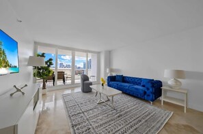 A modern living room with a blue sofa, white coffee table, and a city view through large windows.