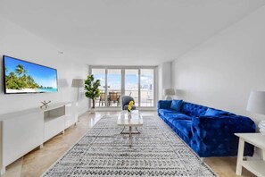 A modern living room with a flat-screen TV, a blue velvet sofa, a coffee table, and a patterned rug.
