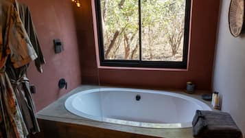 A bathroom with a large bathtub, a window with a view of trees, a towel, and a bottle of soap.
