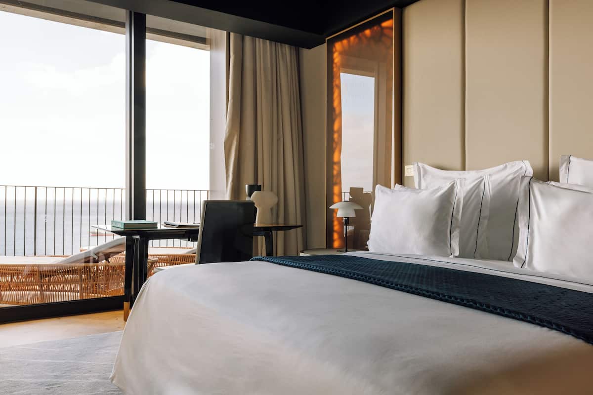 Quarto de hotel com cama grande, uma cadeira, uma mesa pequena e vista para o oceano.