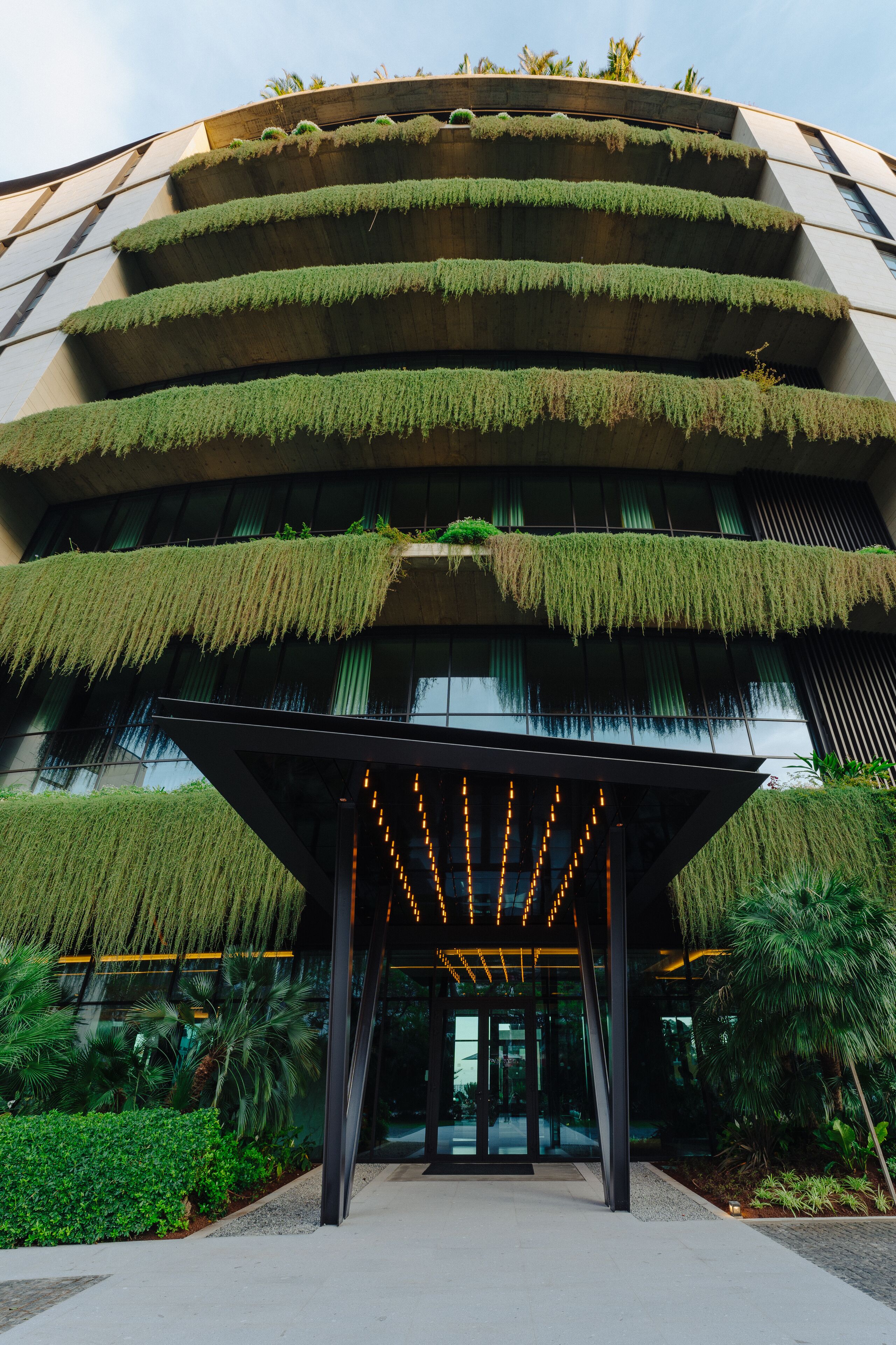 Een modern gebouw met meerdere balkons begroeid met planten, een glazen ingang en een verzorgde tuin eromheen.