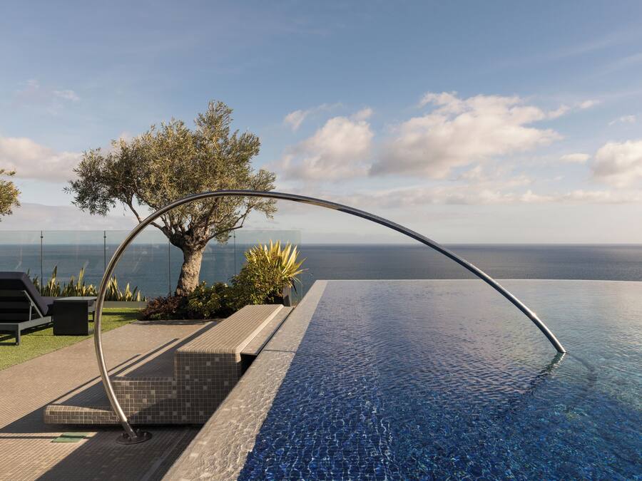 A modern infinity pool with a clear view of the ocean, a tree, and a lounge area with chairs and a small table.