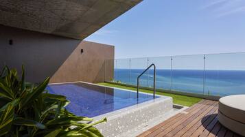 A rooftop pool with a clear view of the ocean, surrounded by wooden decking and potted plants.