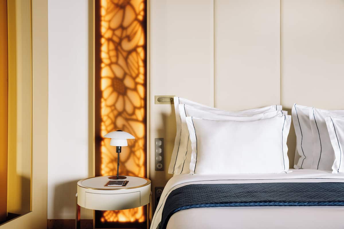 Cama bem arrumada com lençóis brancos e um toque de azul, uma mesinha de cabeceira com abajur e uma parede com painel decorativo.