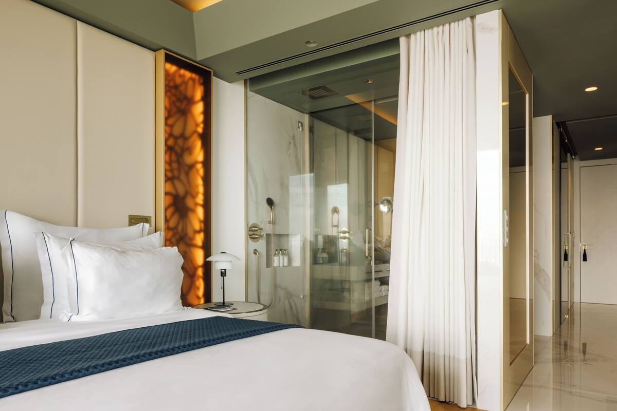 Quarto de hotel moderno com uma cama grande, box de vidro para o chuveiro e vista da cidade.