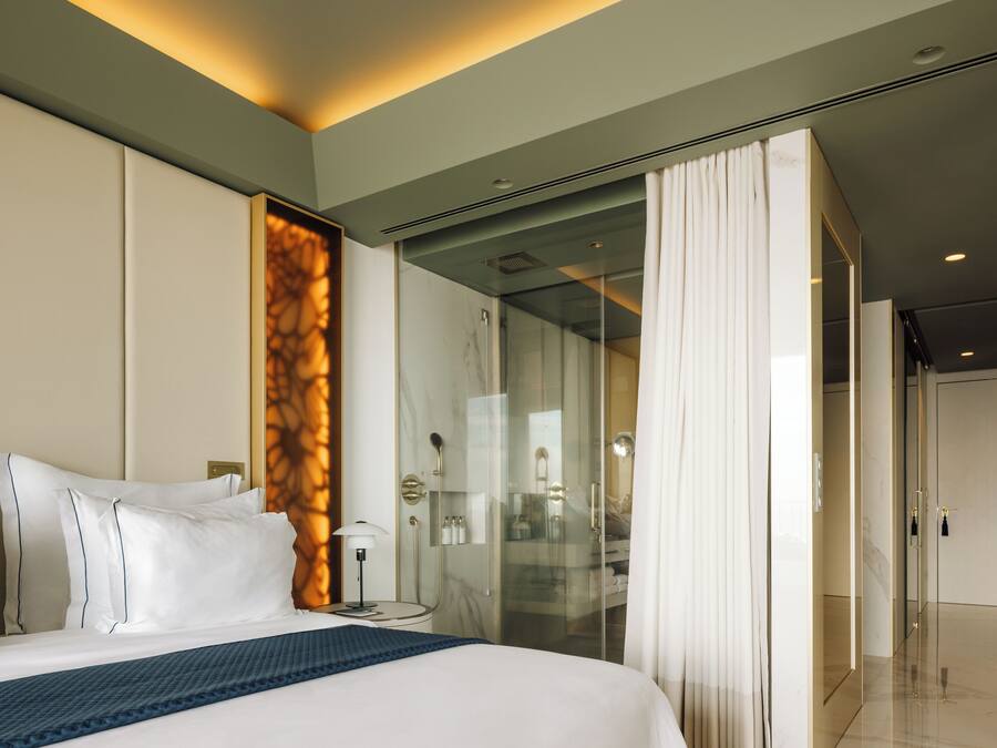 A modern hotel room with a large bed, a glass shower enclosure, and a view of the city.