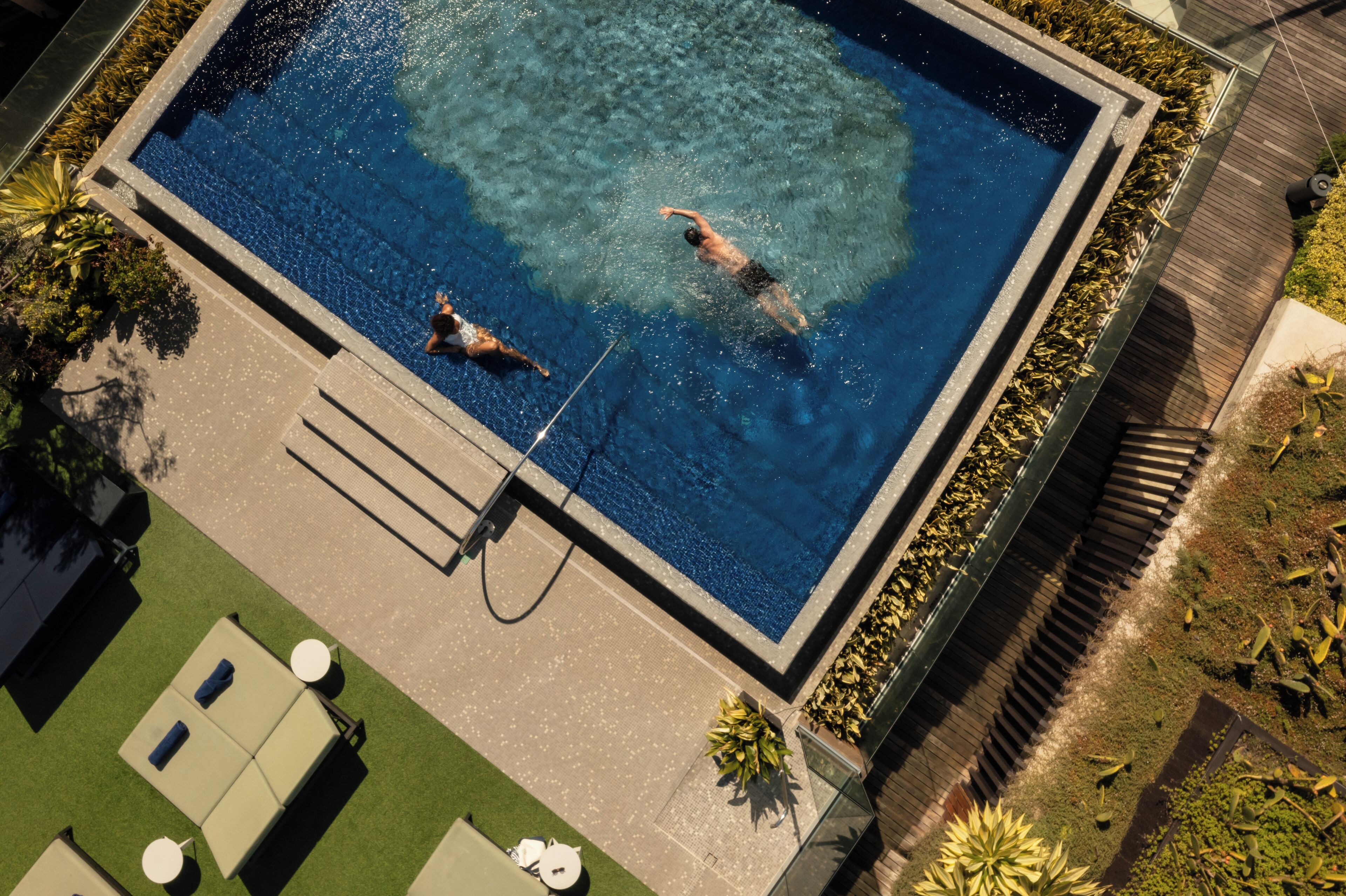 An aerial view of a rectangular swimming pool with two people swimming, surrounded by a deck and greenery.