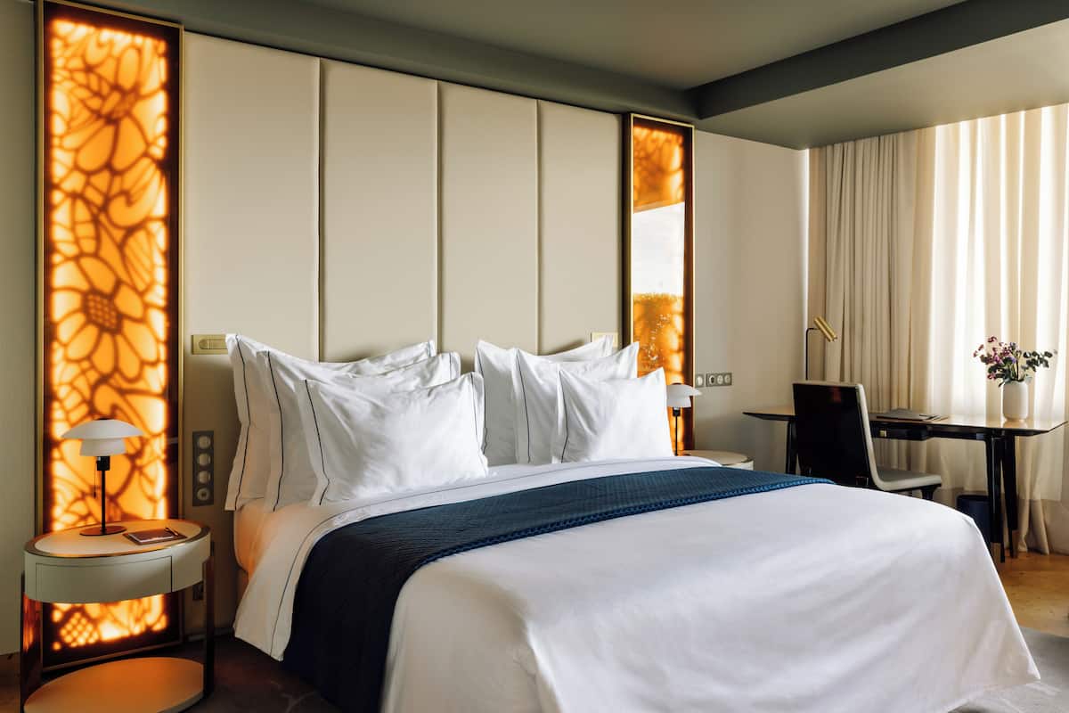 Quarto de hotel com uma cama grande, uma escrivaninha e uma cadeira. Há uma luminária na mesinha de cabeceira e um vaso com flores na escrivaninha.