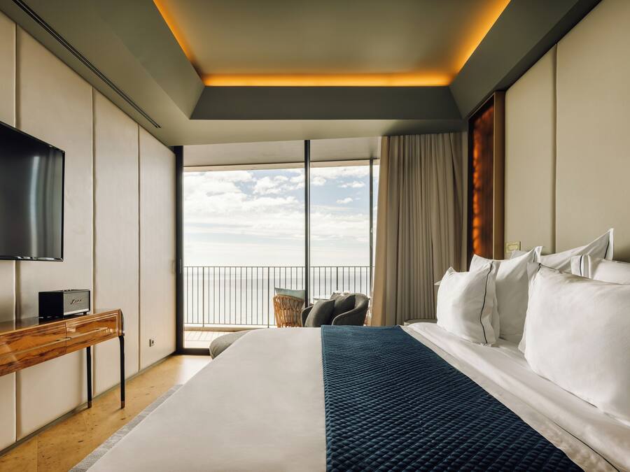 A modern hotel room with a large bed, a flat-screen TV, a wooden desk, and a balcony with a view of the sky.