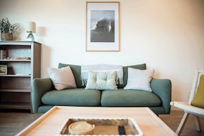 Un divano verde con cuscini, un vassoio di legno con un telecomando, una poltrona in vimini e un quadro appeso al muro.