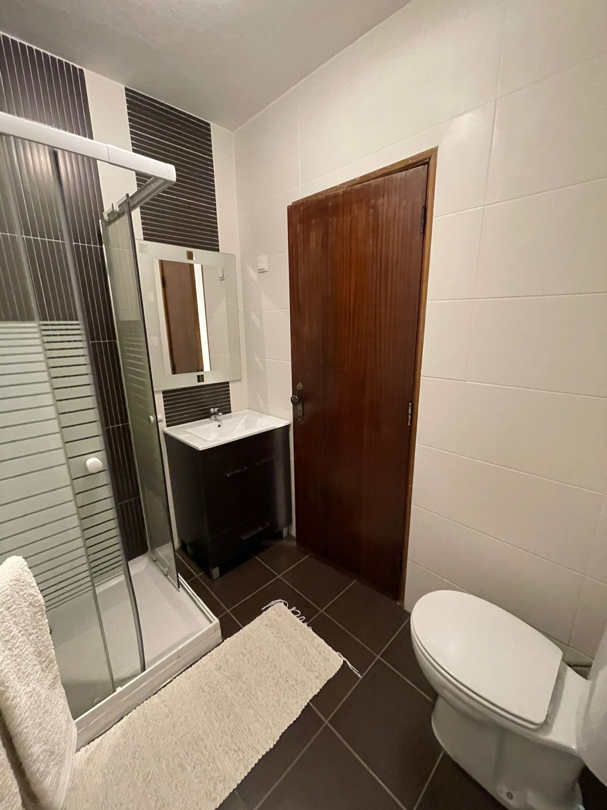 A bathroom with a glass shower enclosure, a wooden door, a white toilet, and a dark-colored sink area.
