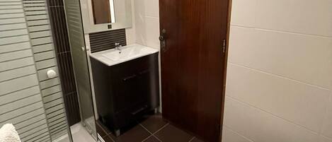 A bathroom with a glass shower enclosure, a wooden door, a white toilet, and a dark-colored sink area.