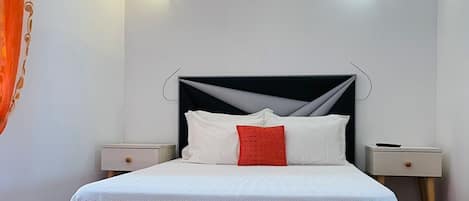 A neatly made bed with white bedding and a red accent pillow, flanked by two bedside tables with lamps, in a room with white walls and a tiled floor.