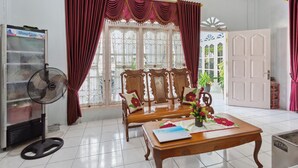 Ein Zimmer mit einer Holz-Sitzecke, einem Tisch mit einem Laptop und Blumen, einem Ventilator, einem Kühlschrank und einem Fenster mit Vorhängen.