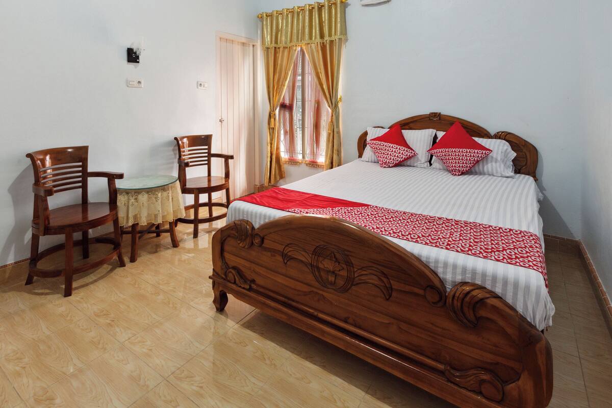 Una cama de madera con colcha roja y blanca, dos sillas de madera, una mesita redonda y un ventanal con cortinas.