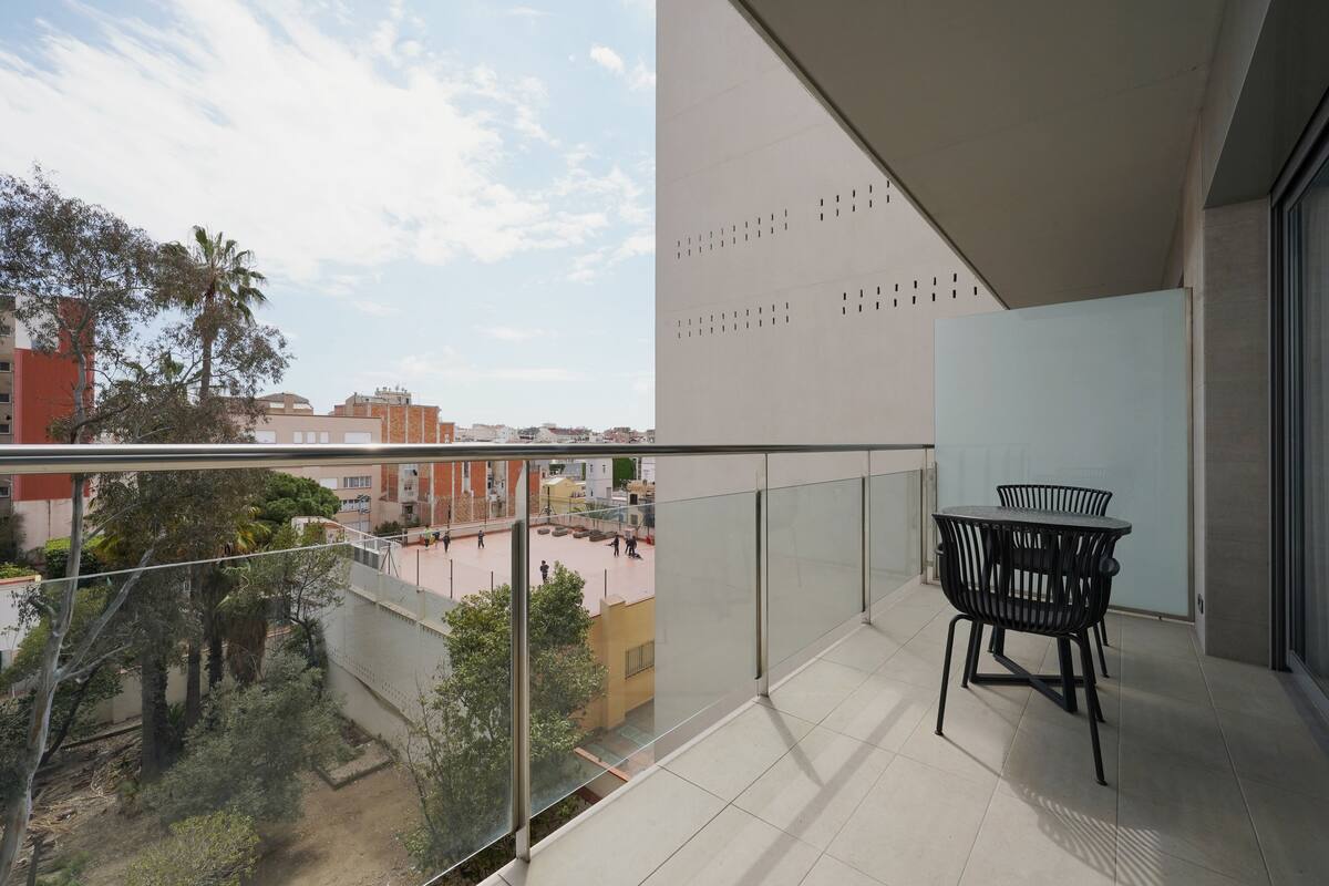 Uma varanda com guarda-corpo de vidro, uma mesa com cadeiras e vista para a paisagem urbana.