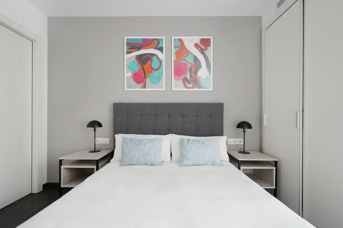 Quarto de hotel moderno com uma cama grande, duas mesas de cabeceira e arte abstrata na parede.