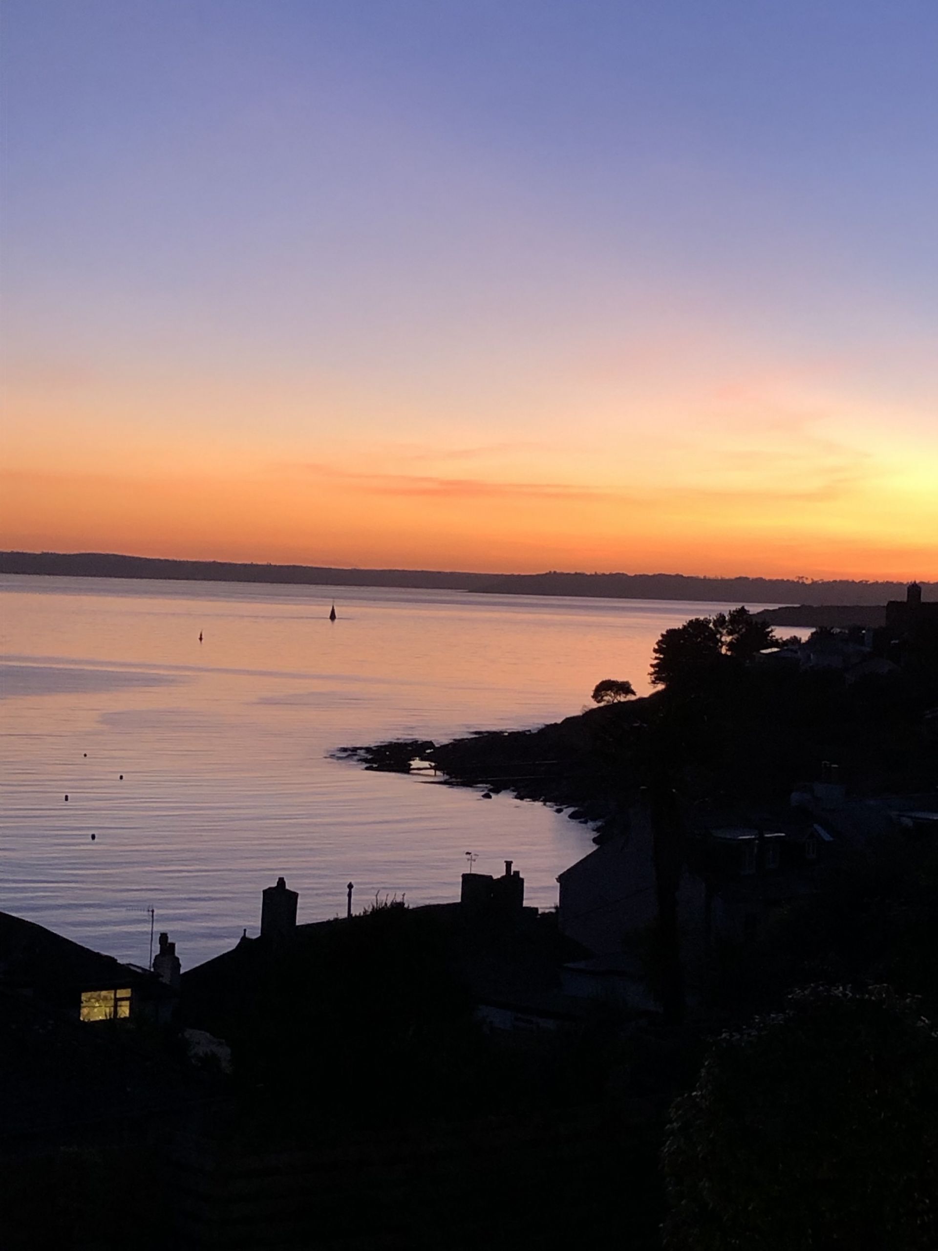 A coastal sunset view with calm waters, a distant shoreline, and silhouetted buildings.