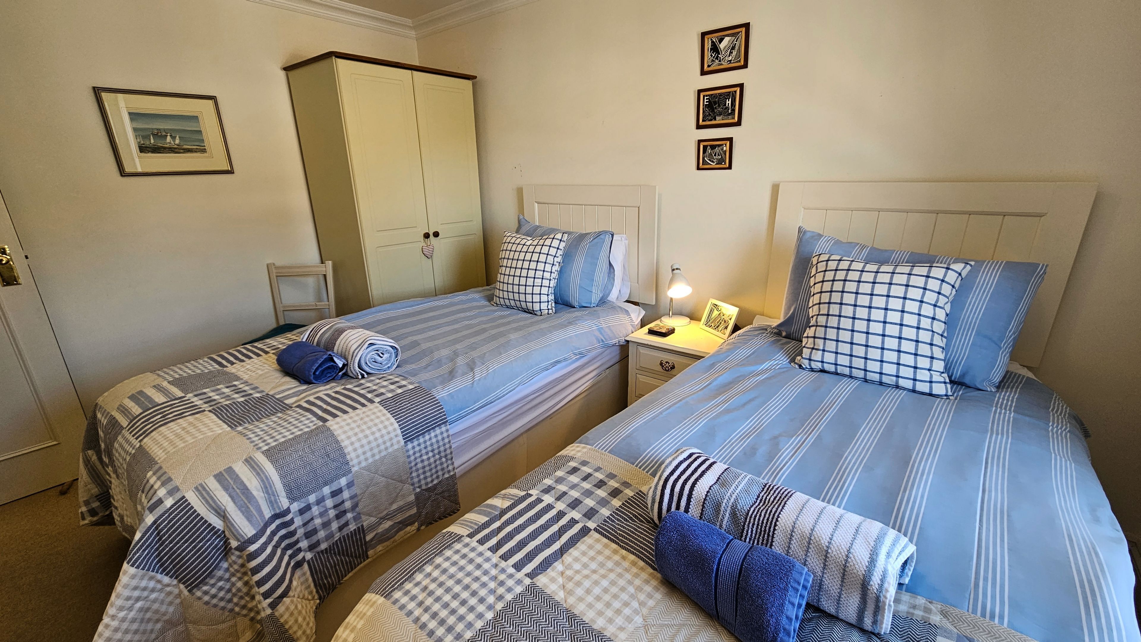 A room with two beds, each with a blue and white striped bedspread and matching pillows. There is a wardrobe, a nightstand with a lamp, and a framed picture on the wall.
