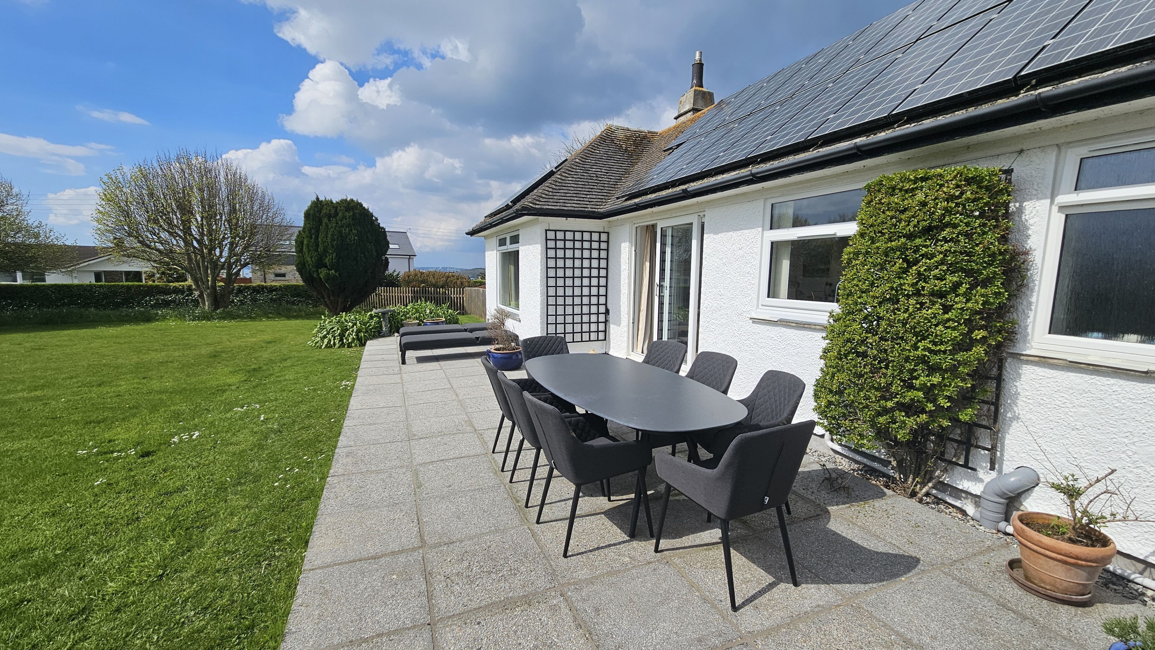 Une terrasse avec une table ronde et des chaises, une maison blanche avec un toit en tuiles et une pelouse verte.