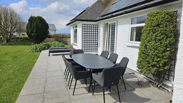 A patio with a table and chairs, a white building with a black gate, and a hedge.