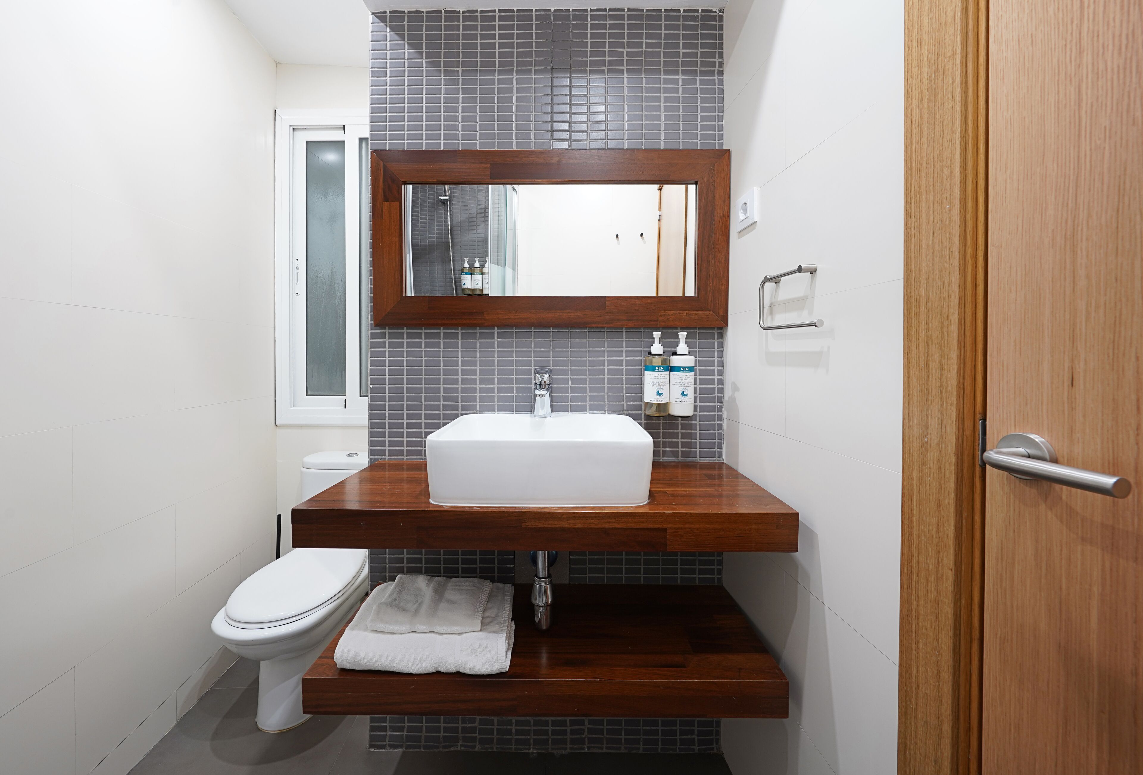Un baño con inodoro blanco, tocador de madera con lavamanos blanco y un espejo sobre el lavamanos.