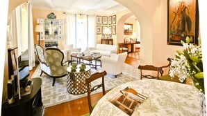 A spacious living room with a white sofa, a coffee table, and a dining table with a patterned tablecloth. There are chairs, a bookshelf, and a painting on the wall.