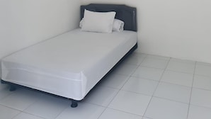 A small, single-bed room with a white mattress, a dark headboard, and a white wall. There is a white air conditioning unit mounted on the wall above the bed.