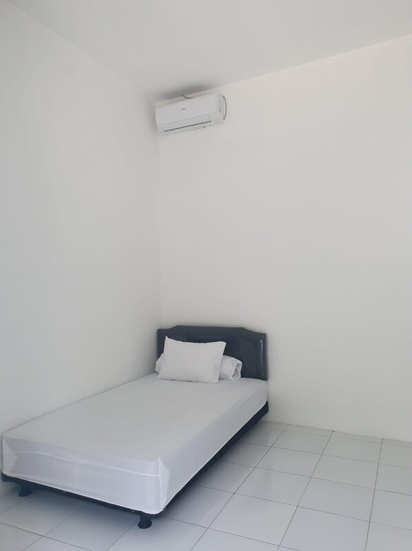 Una habitación pequeña con cama individual, colchón blanco, cabecera oscura y pared blanca. Se observa un equipo de aire acondicionado blanco instalado en la pared sobre la cama.