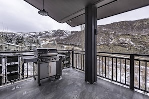 A balcony with a grill and outdoor seating, overlooking a snowy mountainous landscape.