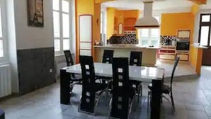 Un coin repas avec une table blanche et des chaises noires, une cuisine en arrière-plan et une fenêtre laissant entrer la lumière naturelle.