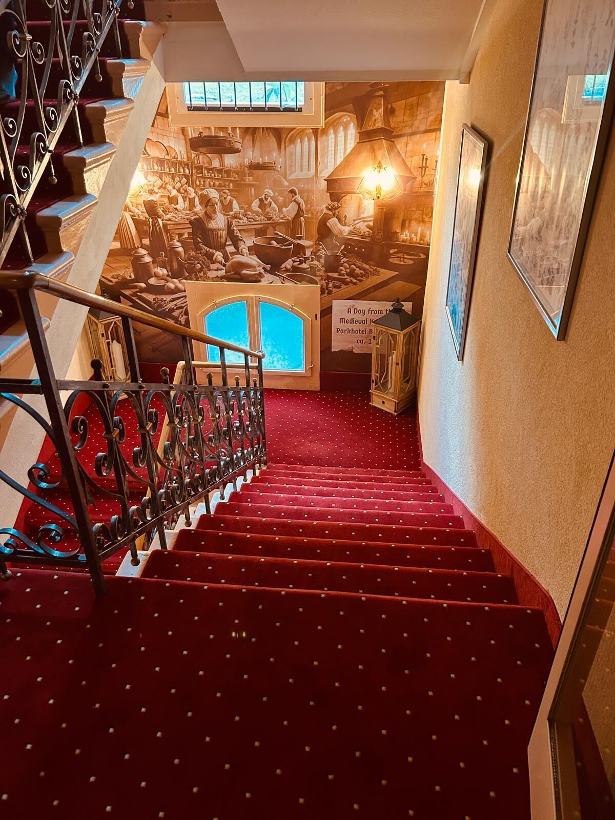 Un escalier recouvert d’une moquette rouge et doté de balustrades en métal mène à une pièce ornée d’une fresque murale sur le thème du Moyen Âge.