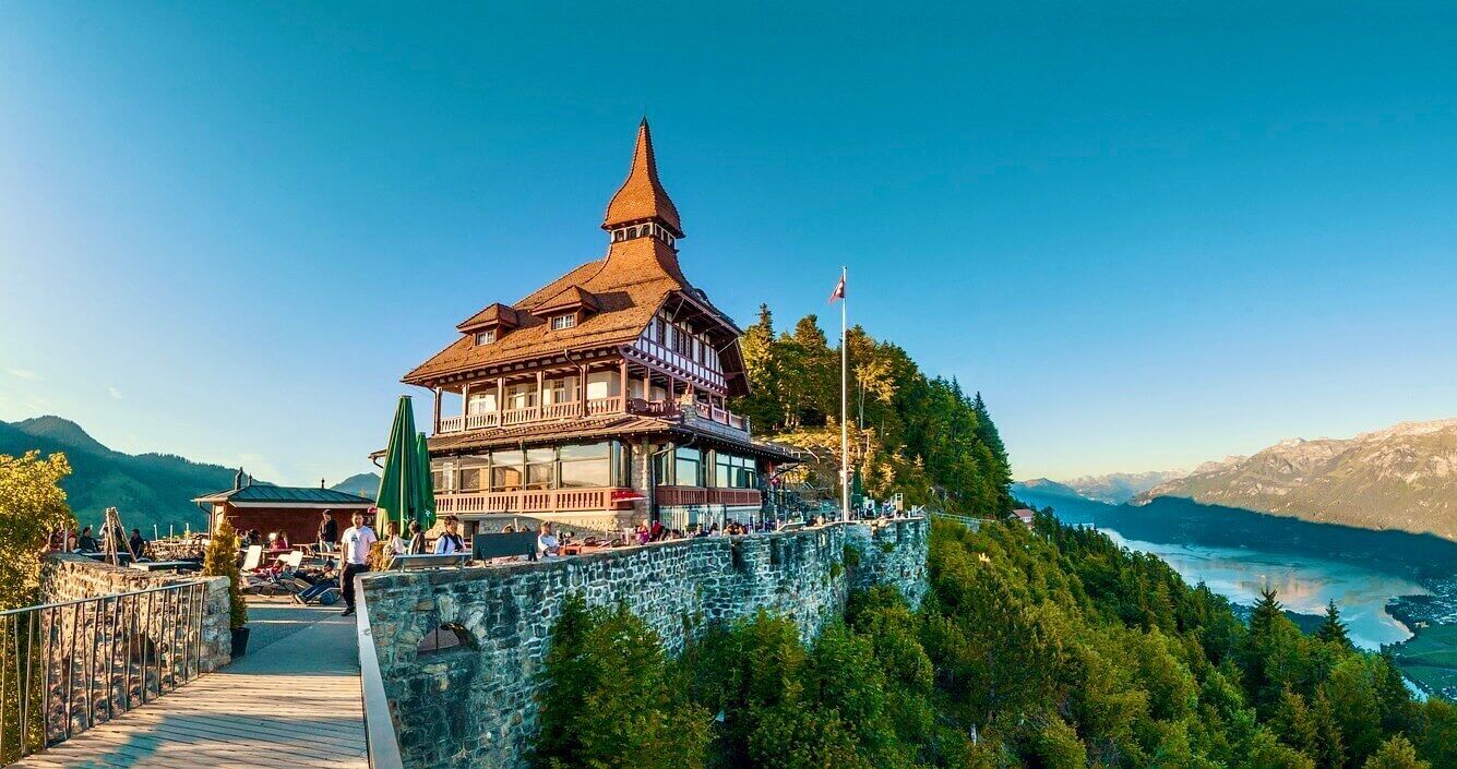 Il s’agit d’un bâtiment de style chalet traditionnel, doté d’un toit à forte pente et d’une tour, situé sur un mur de pierre surplombant un paysage montagneux et un lac.