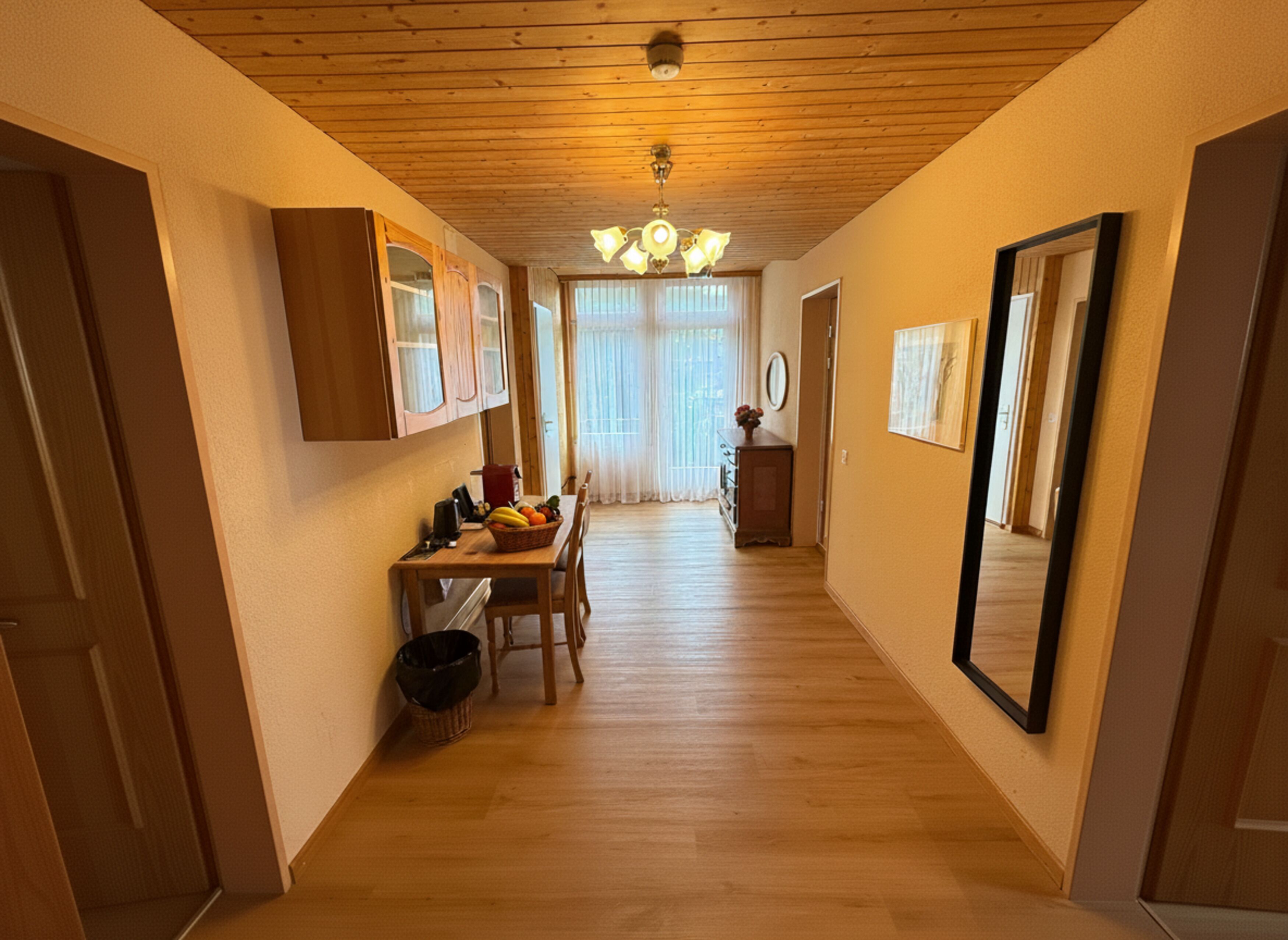 Un pasillo con suelo de madera, una zona de comedor con mesa y sillas, y un espejo en la pared derecha.