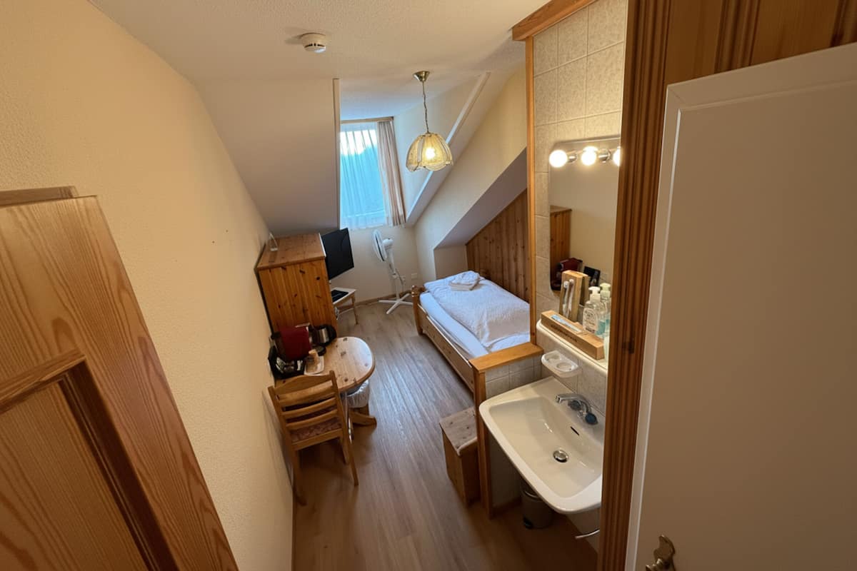 Habitación compacta con cama, escritorio, silla, mesita, lavamanos y espejo.