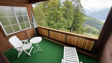 Un balcón con piso de madera, dos sillas blancas, una mesita y vistas a un paisaje verde con árboles y montañas.