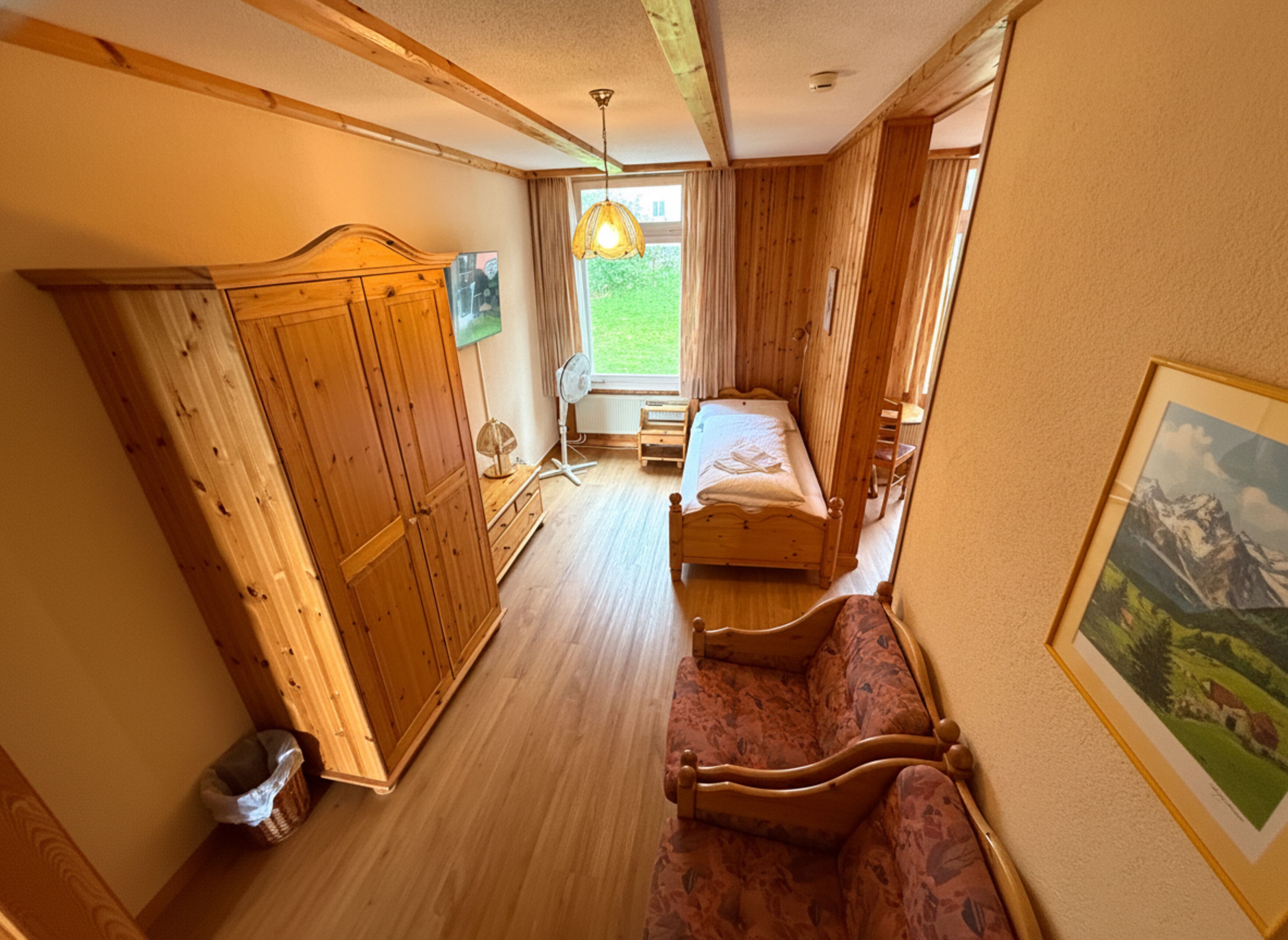 Ein Zimmer mit Holzschrank, Bett, Stuhl und einem Fenster mit Blick ins Grüne.
