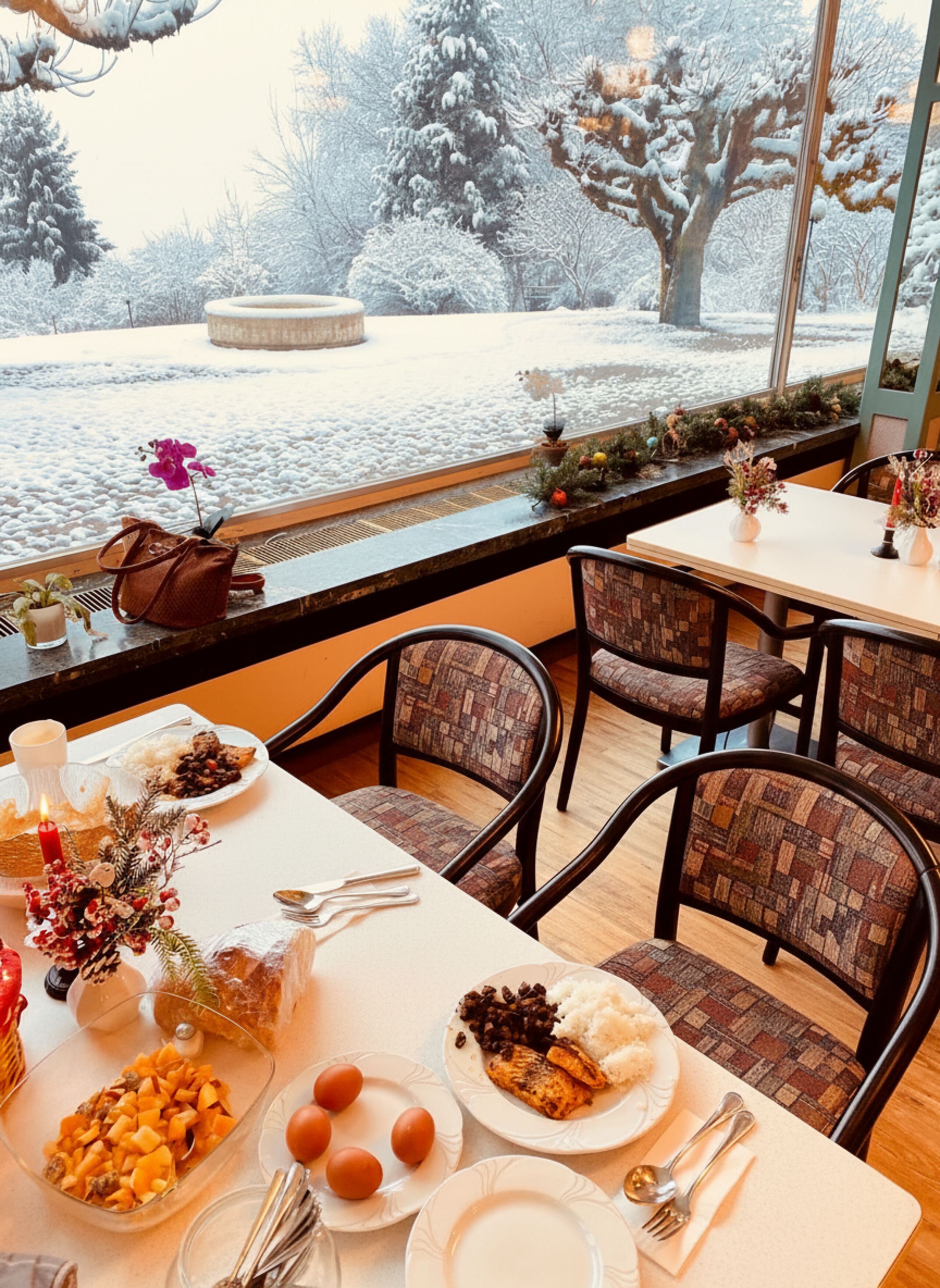 A dining area with a table set for two, featuring a meal with rice, beans, eggs, and a piece of grilled meat. There are chairs, a vase with flowers, and a view of a snowy landscape through a window.