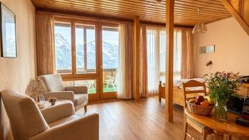 Ein gemütliches Zimmer mit Holzbalkendecke, einer Couch, einem Esstisch mit Blumenvase und Bergblick durch große Fenster.