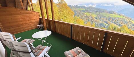 A balcony with a green carpet, white chairs, a small round table, and a patterned sofa.