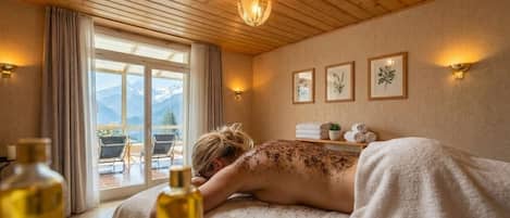 Une salle de spa avec un plafond en bois, une table de massage, une fenêtre donnant sur les montagnes et des reproductions encadrées de gravures botaniques au mur.
